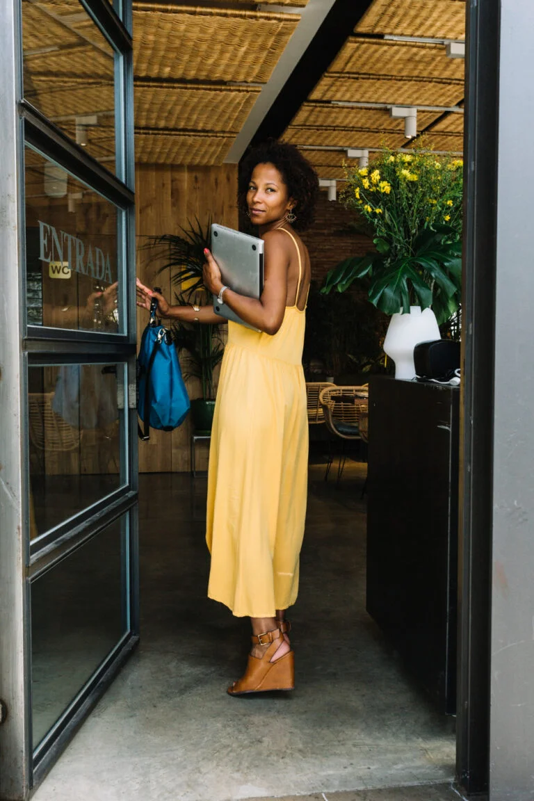 Moça segurando a porta da entrada de um coworking. Ela está de vestido longo amarelo e segurando um notebook em uma das mãos