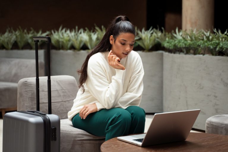 mulher ansiosa pela sua viagem a trabalho, ela está pensativa olhando para o notebook e próxima da mala de viagem