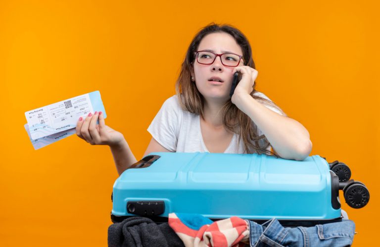 Mulher ao telefone segurando duas passagens na outra mão e apoiada sobre a mala preocupada com seu passaporte