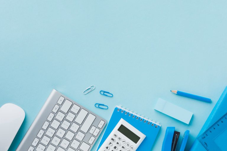 Mesa de trabalho organizada em tons de azul, com teclado, mouse, calculadora, bloco de anotações, lápis, clipes de papel, régua e grampeador sobre fundo azul-claro, transmitindo sensação de ordem e produtividade.